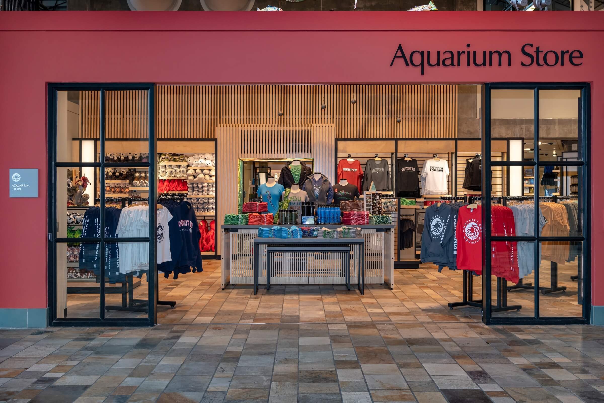A view of the entrance of the Aquarium store from the main lobby of the Aquarium. Various apparel, including logo shirts, jackets,and plushes sit on hangers and shelves