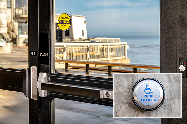 Glass doors leading to the back deck at the Aquarium; one says push while the other says pull. An inset in the bottom right corner shows a closeup of the silver button, with the blue international symbol of access above blue text that reads ‘push to open’
