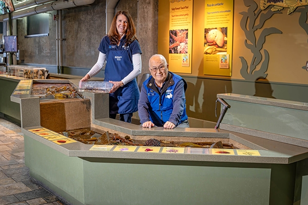 Two Aquarium volunteers in blue welcome you to the lower section of the touch pool. One volunteer is sitting behind the lower area, while another stands off behind him, holding a clear acrylic tray that can be used to bring animals closer to guests in wheelchairs