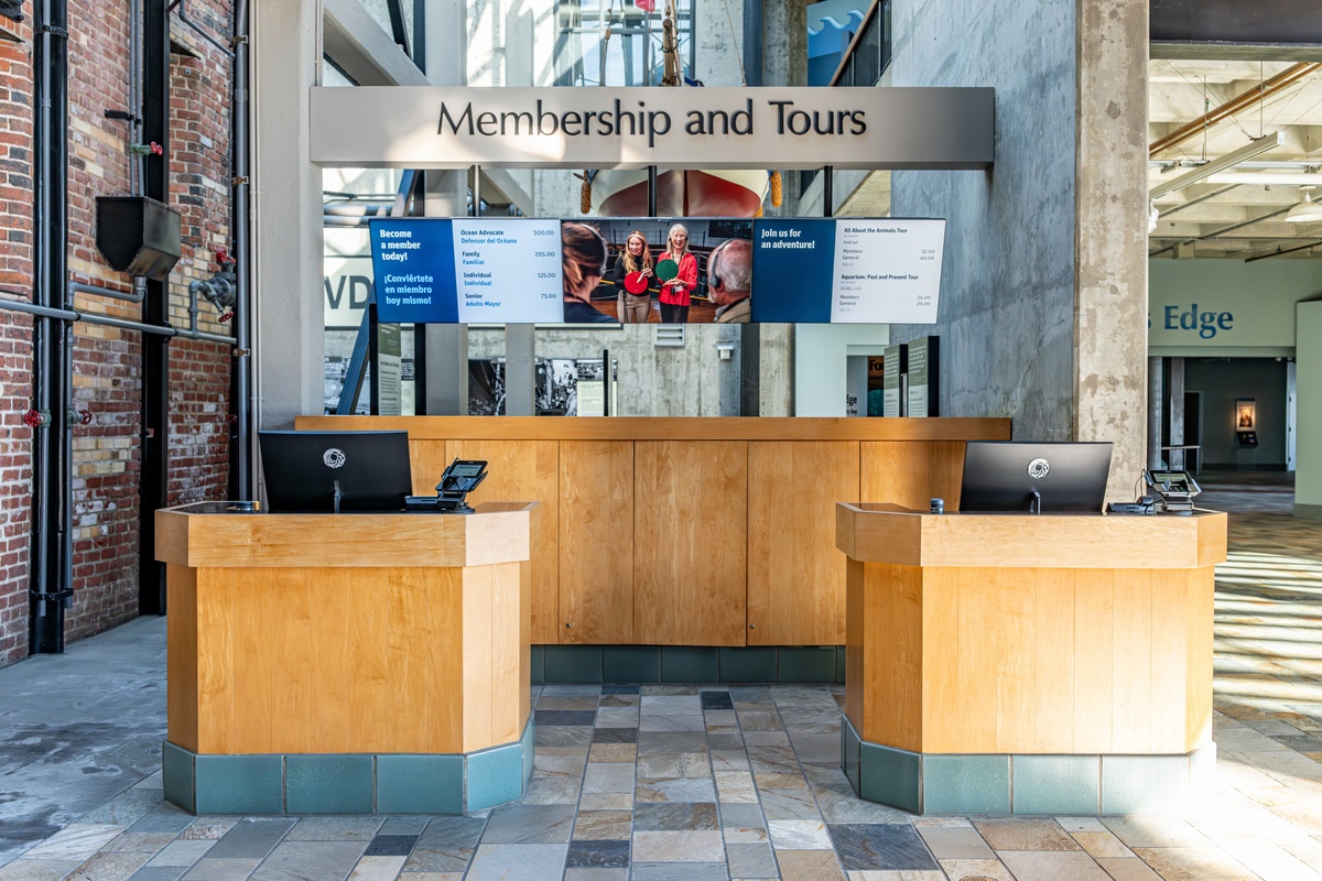 Two wooden kiosks stand in front of a larger wooden cabinet, underneath a series of television screens and a sign that reads, “Membership and Tours”