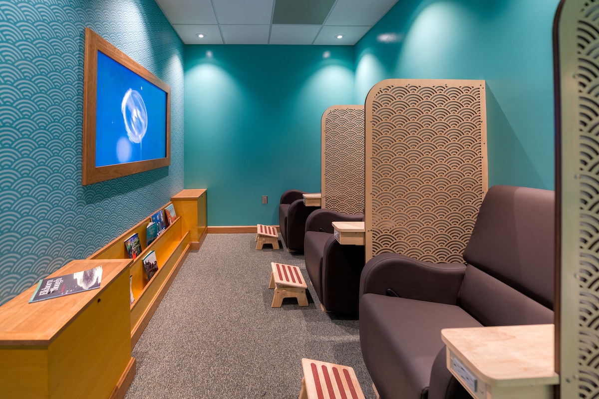The nursing lounge at the Aquarium features teal green walls, a long wooden bookshelf on one side with a wood-framed television above. On the opposite wall are three burgundy lounge chairs with foot rests, with privacy separators in between