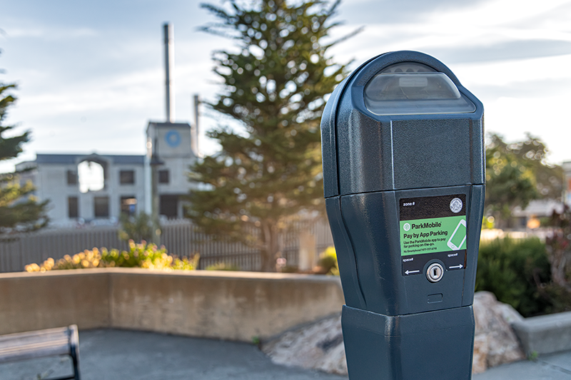 A closeup of a grey parking meter, with the blurred Monterey Bay Aquarium building in the background