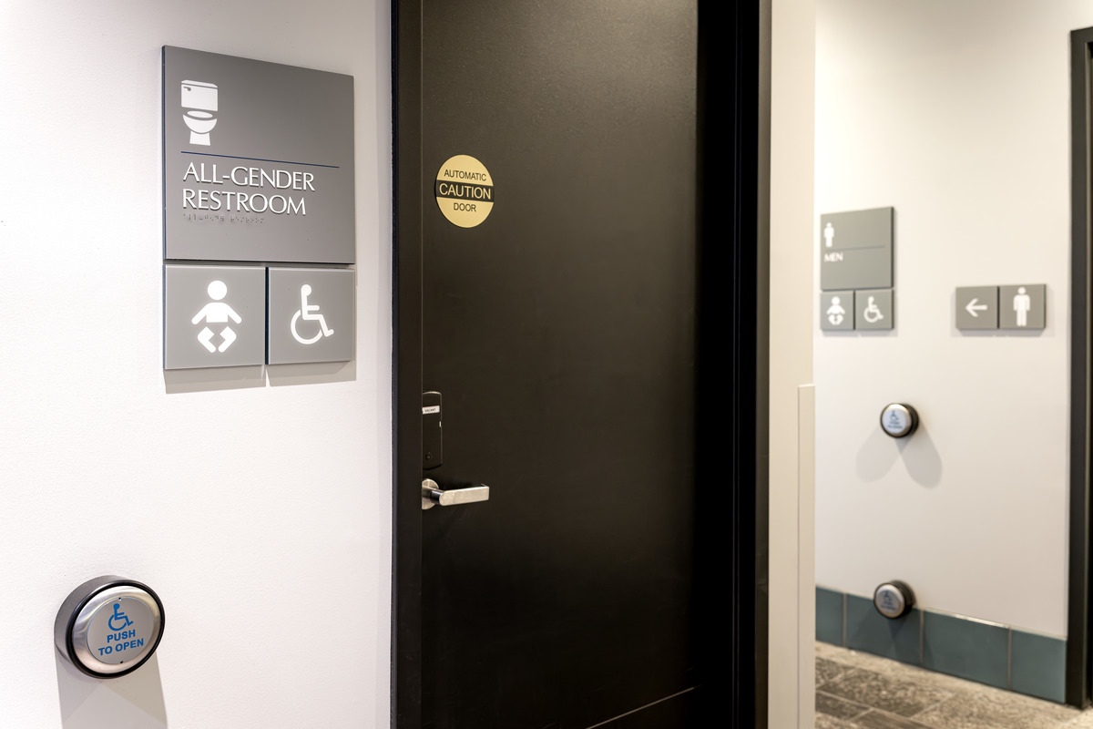 The black door of the all-gender restroom, next to a grey signs denoting an all-gender restroom, handicap accessibility, and a changing table, as well as a silver electronic assist button