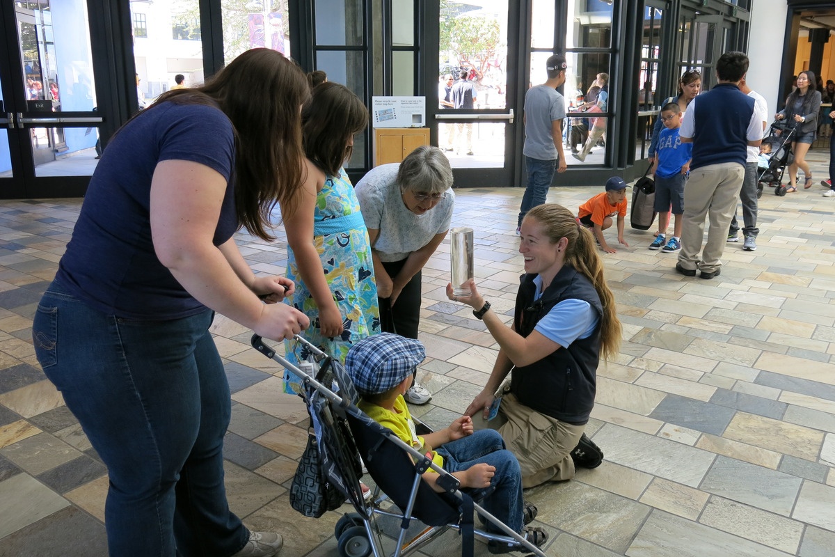 A staff member kneels on the Aquarium floor with a smile on their face while showing a clear tube of moon jellies to a group of people. One of the people is slightly hunched over with their hands on the handles of a stroller with a child in it