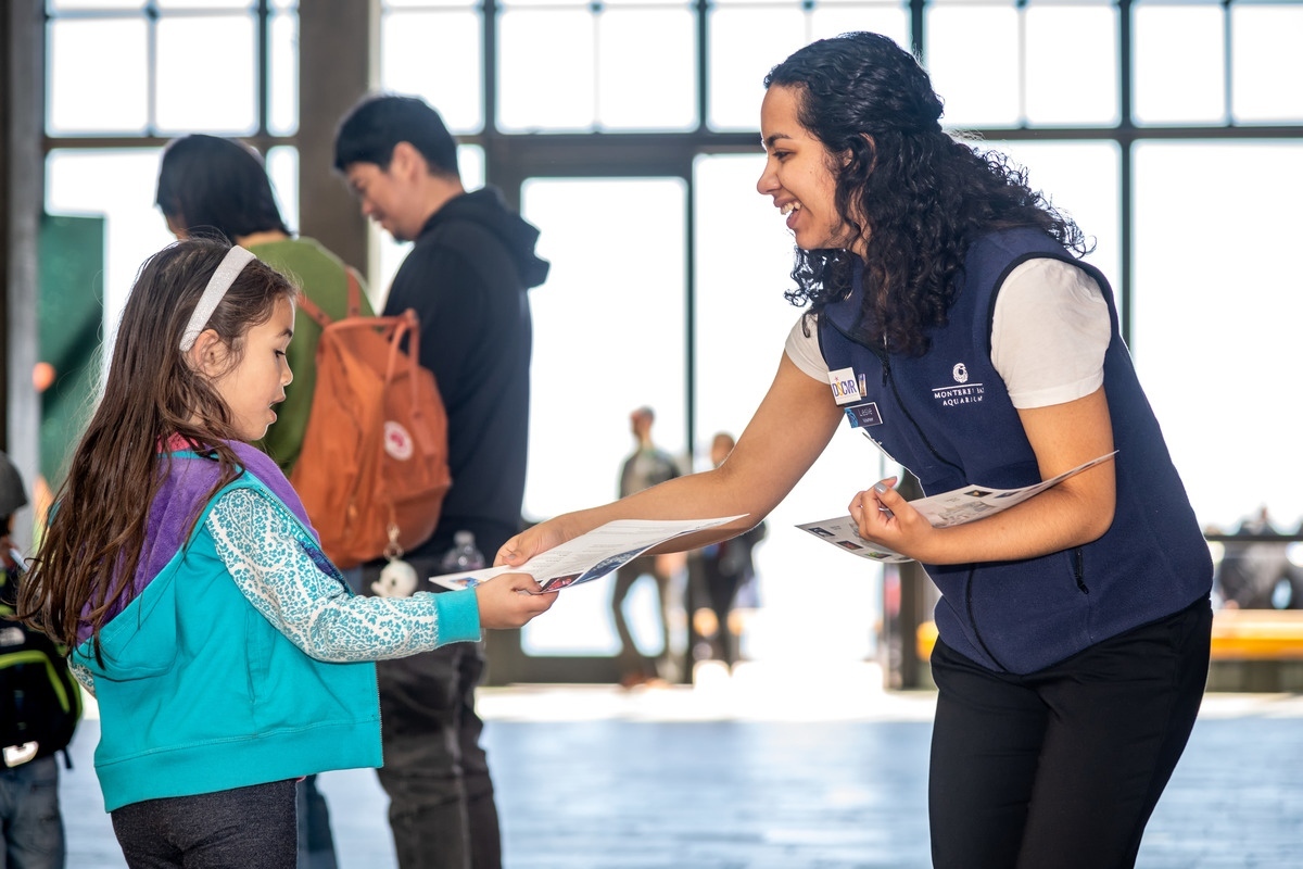A smiling Aquarium volunteer hands a paper program guide to a child