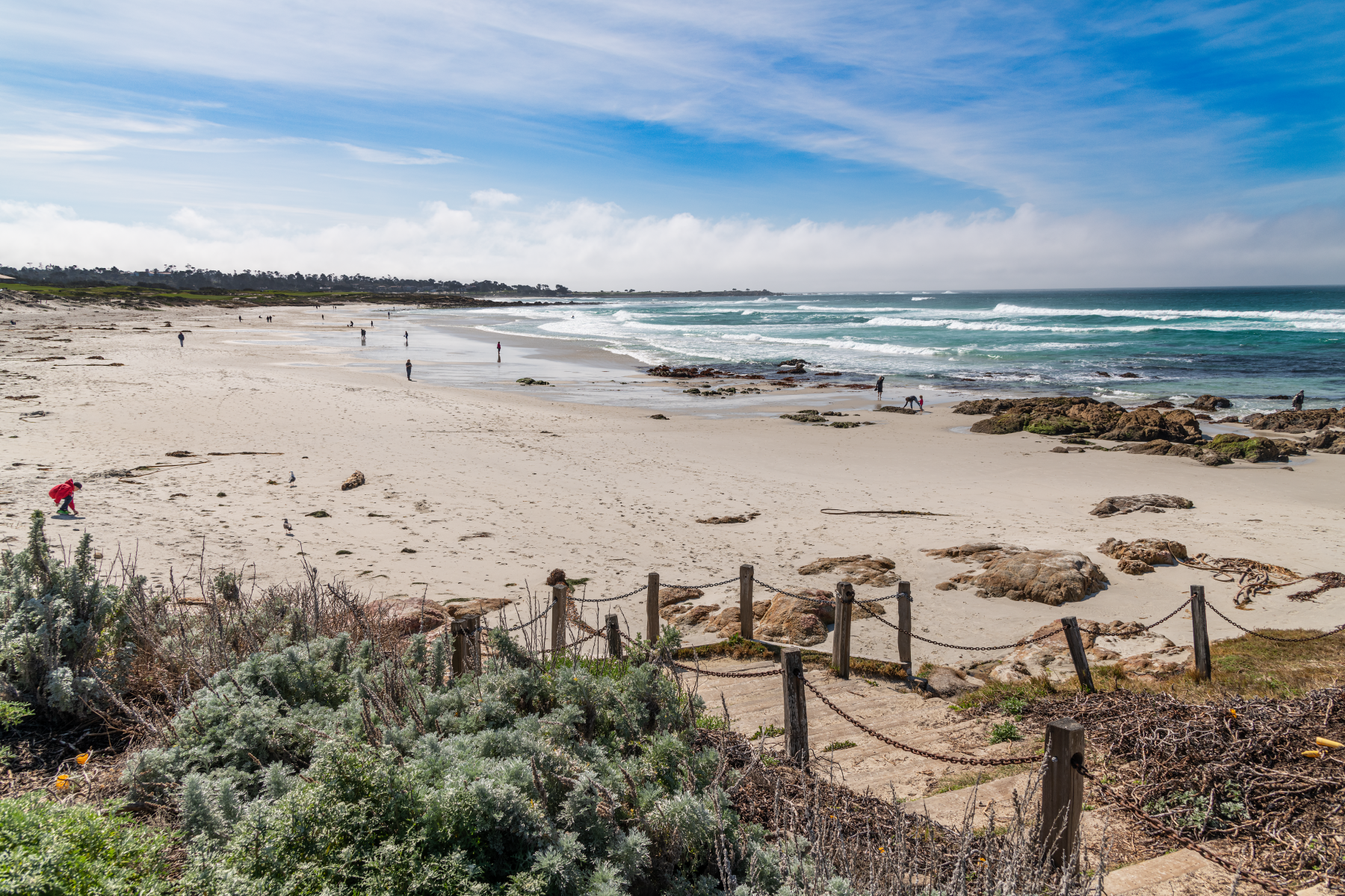 Visitors walking along a wide sandy beach with rocky tide pools waves and coastal shrubs along a chain fence