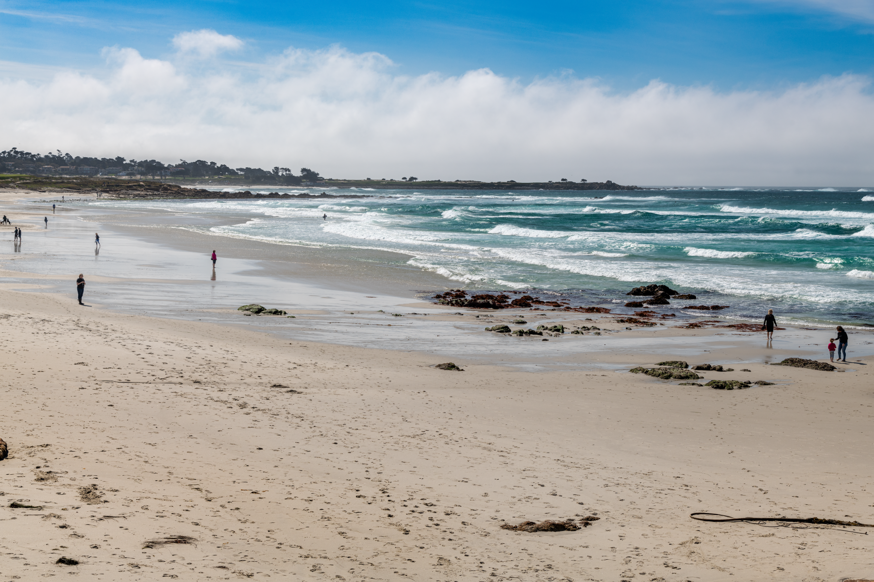 People walking along a wide sandy beach with turquoise waves and rocky outcrops under a partly cloudy sky