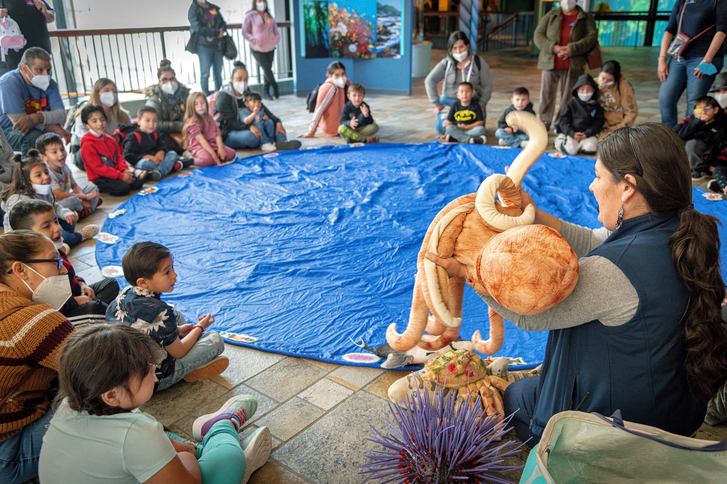 Head Start preschool students and their families gather around a blue parachute on the ground near the Aquarium's Splash Zone exhibit