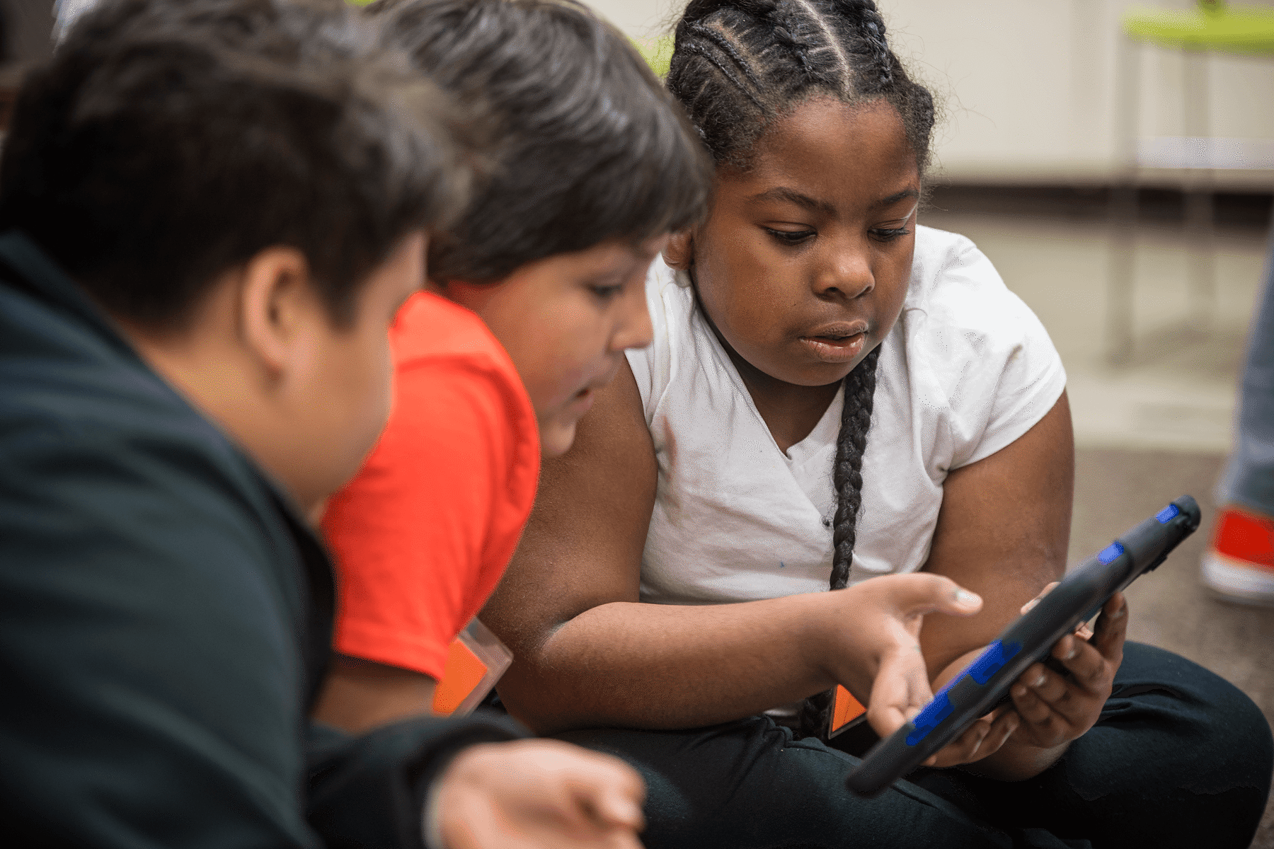 A group of elementary students sit on the floor, leaning in to collaborate on a digital lesson displayed on a tablet held by one of the young students