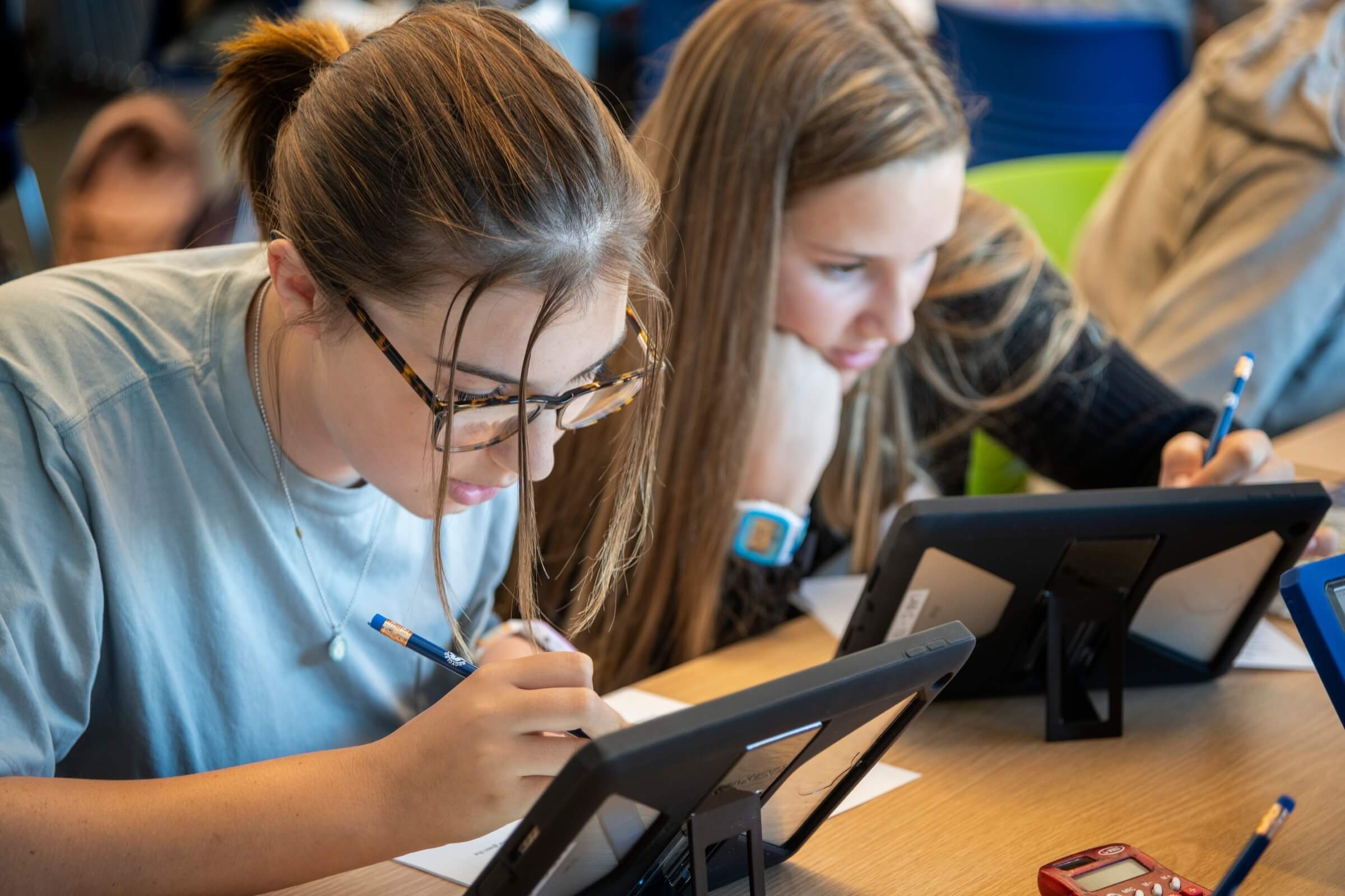 Two students leaning over tablets and writing on paper at a classroom table, focused on their work