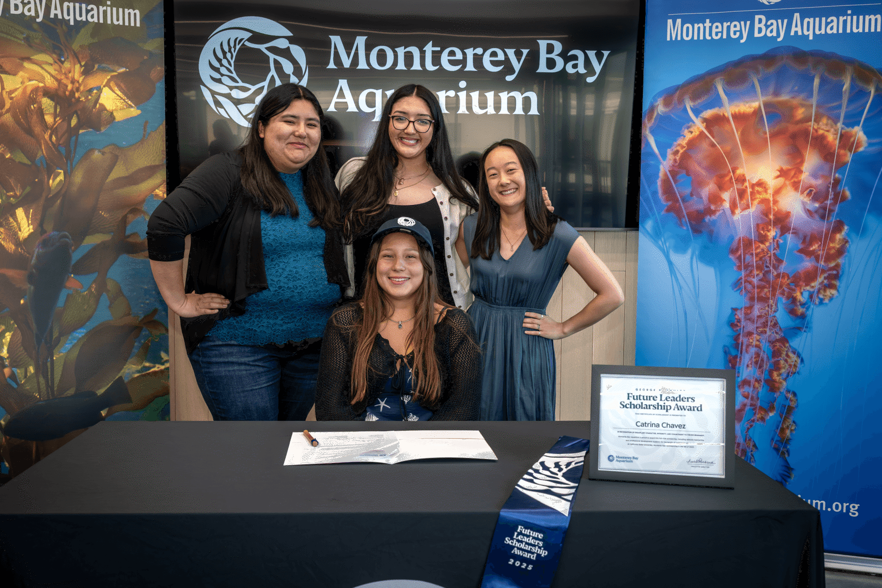 Four smiling teens posing together at a table displaying a Future Leaders Scholarship Award certificate in front of a Monterey Bay Aquarium backdrop