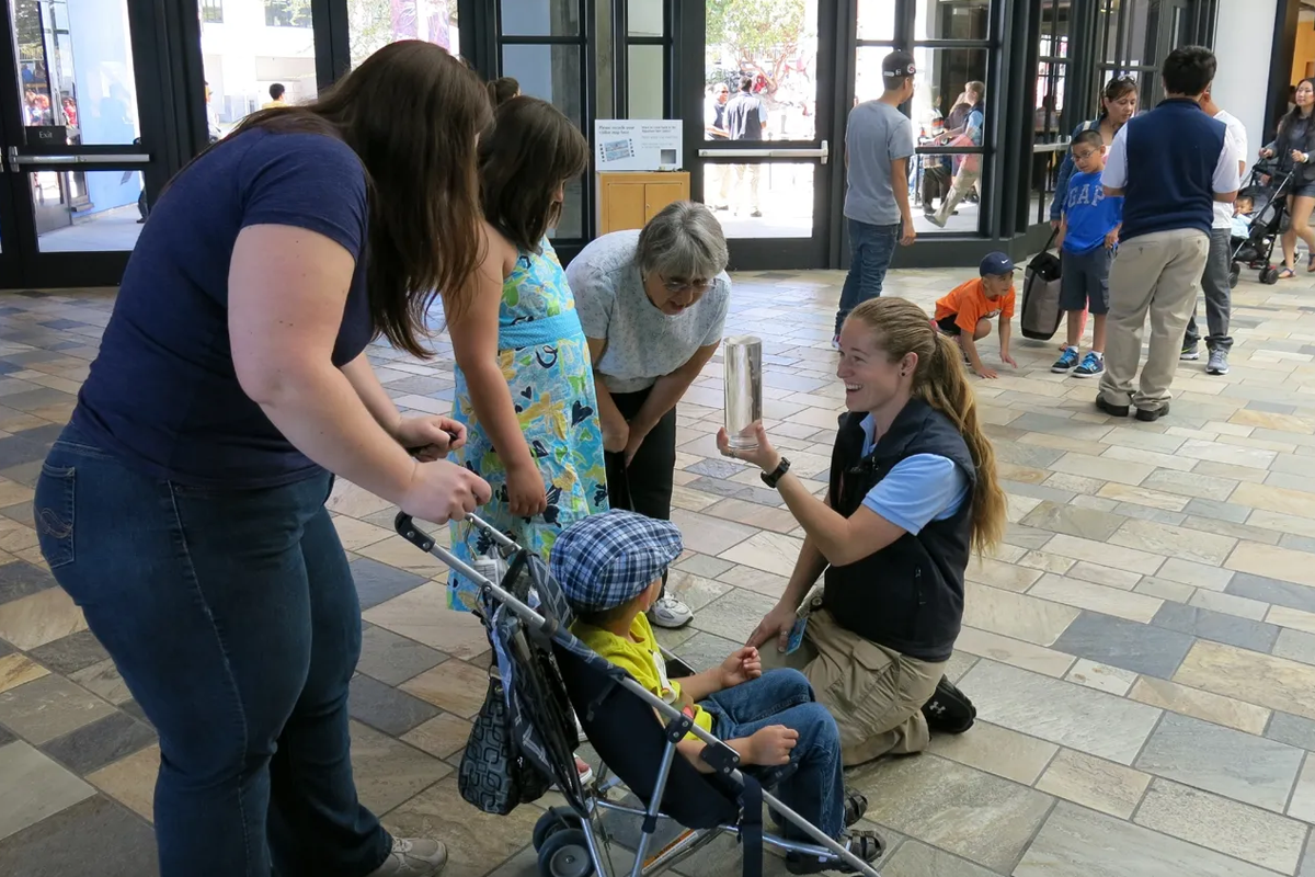 A teen program participant kneeling on the Aquarium floor showing a young visitor in a stroller and their family an up-close view of jellies in a  clear tube