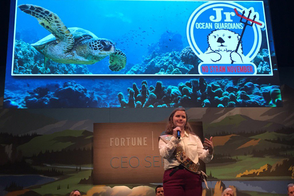 Person holding a microphone and speaking on stage at a conference with a large screen showing a sea turtle and a Jr. Ocean Guardians graphic