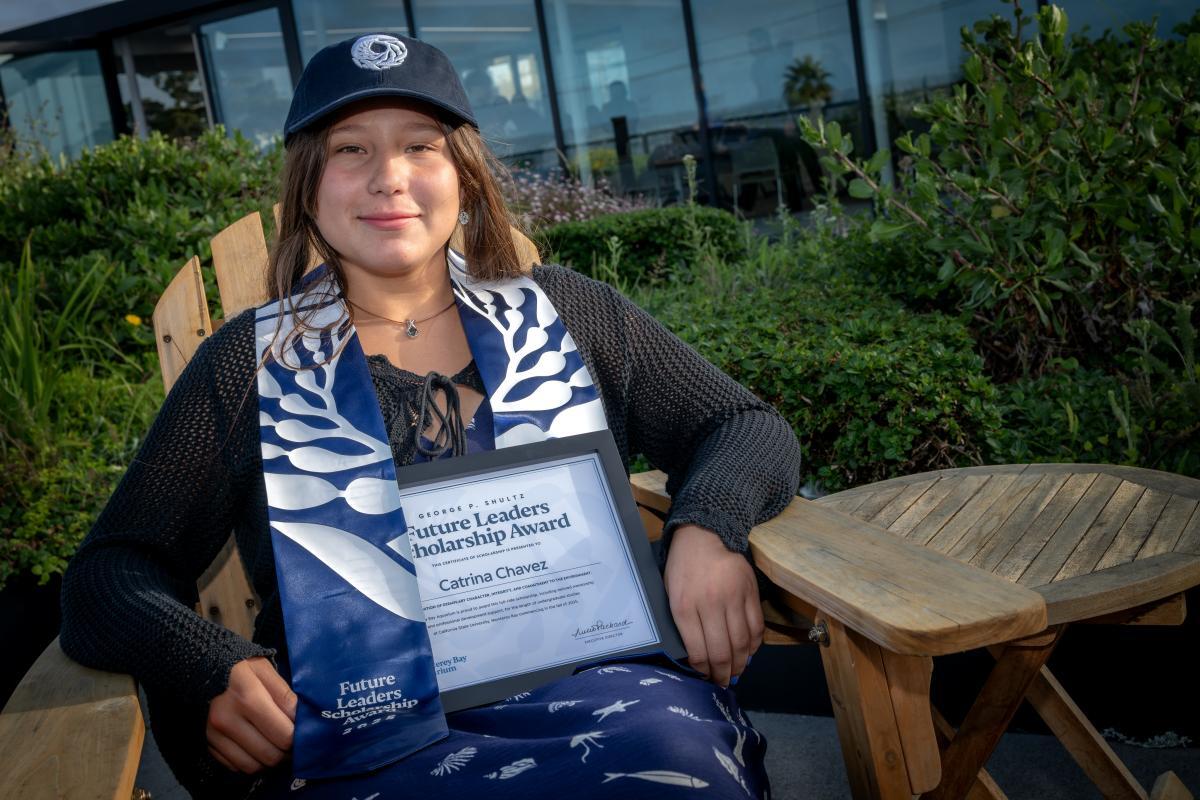 A scholarship recipient sits while wearing a baseball cap and sash with Aquarium logos and her award plaque in her lap