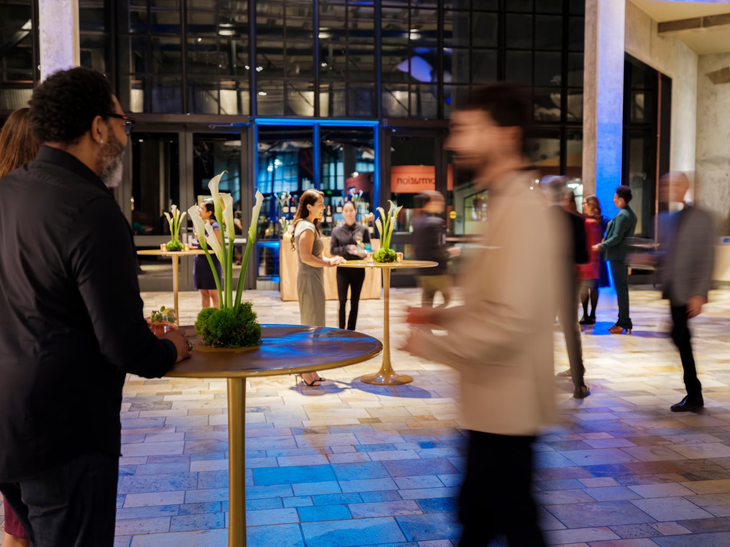 People in business casual attire drinking and chatting around several high-top tables during an evening event at the Monterey Bay Aquarium