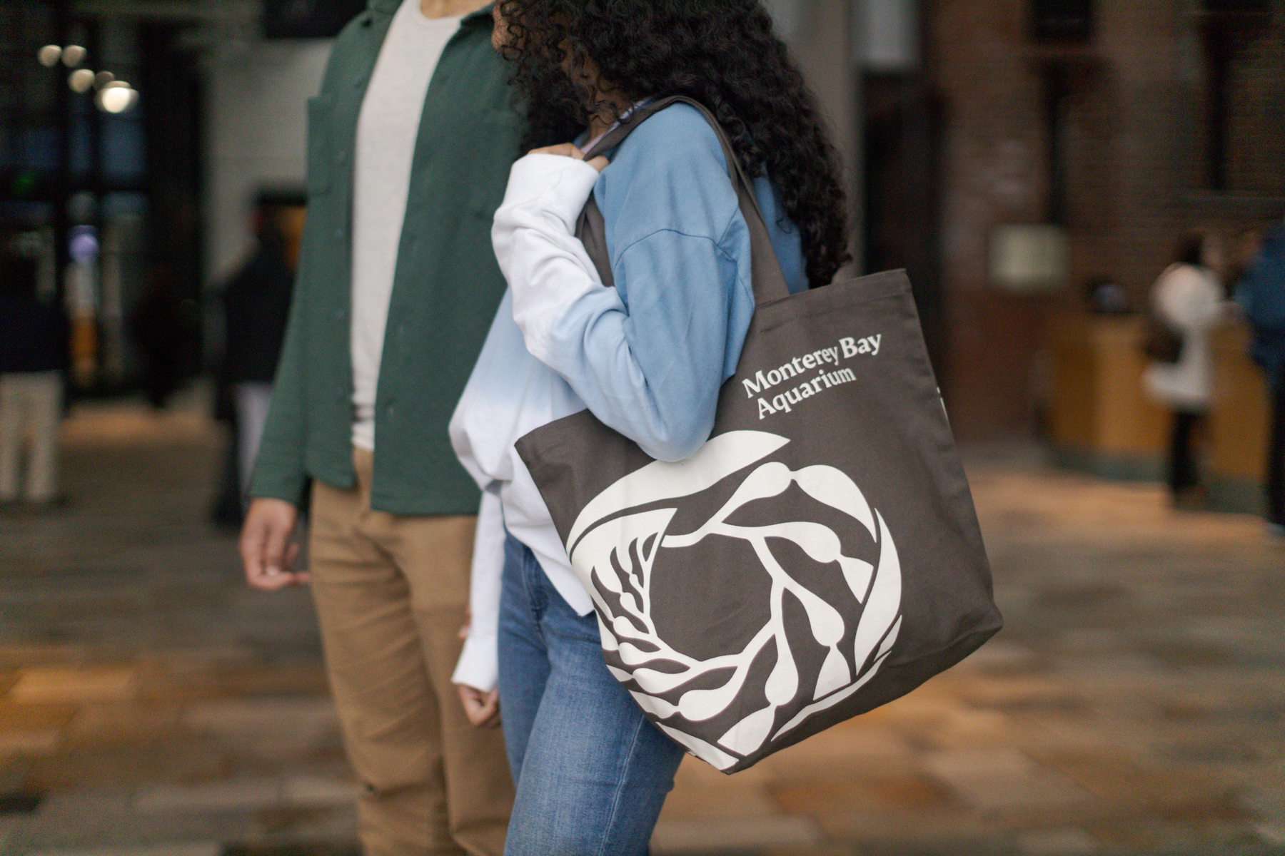 A person carrying a tote bag with “Monterey Bay Aquarium” printed on it walks indoors beside another person.