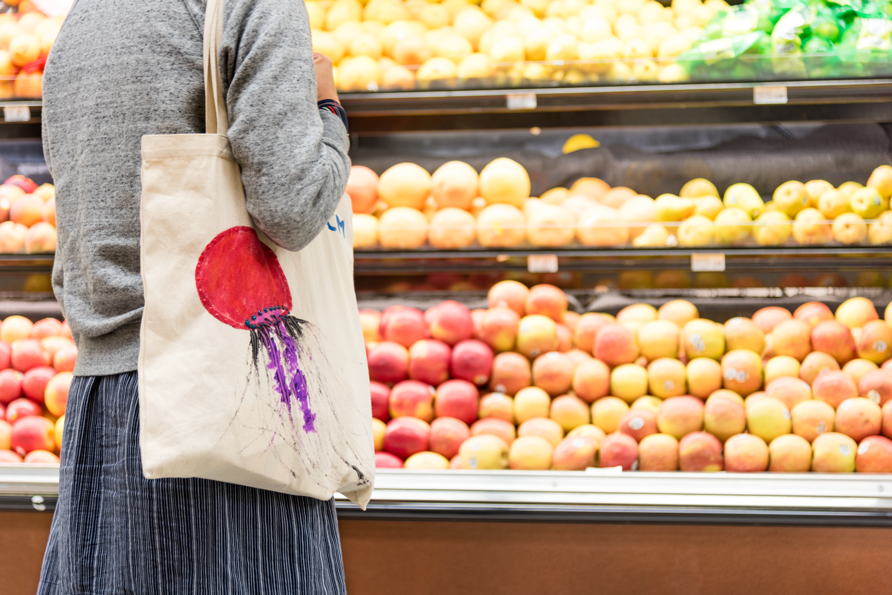 A shopper standing in front of produce at a grocery store holding a reusable bag with a hand painted jellyfish