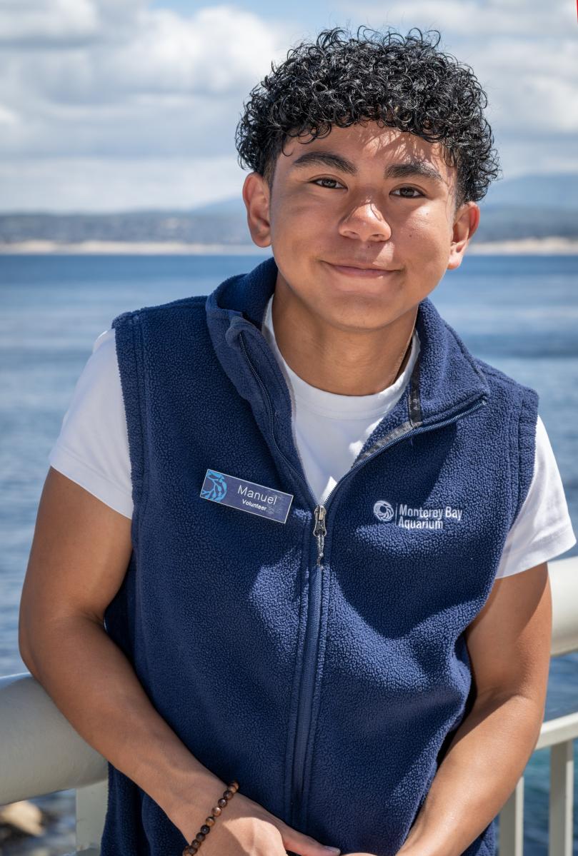 A teen volunteer with short curly black hair and wearing a blue vest stands in front of ocean background