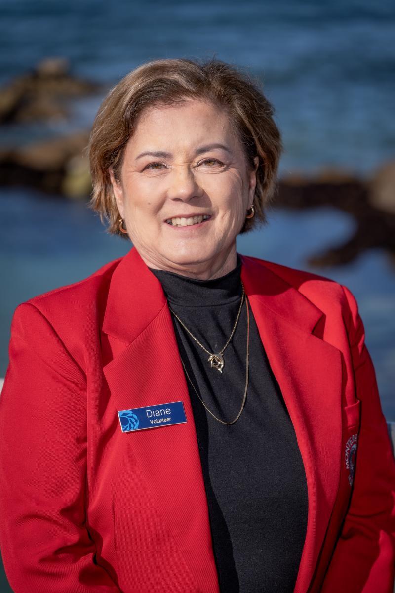 A woman with short brown hair and wearing a red sports jacket smiles against an ocean background