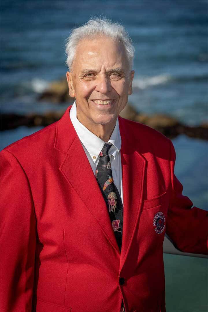 An older gentleman wearing a red sports jacket smiles against an ocean background