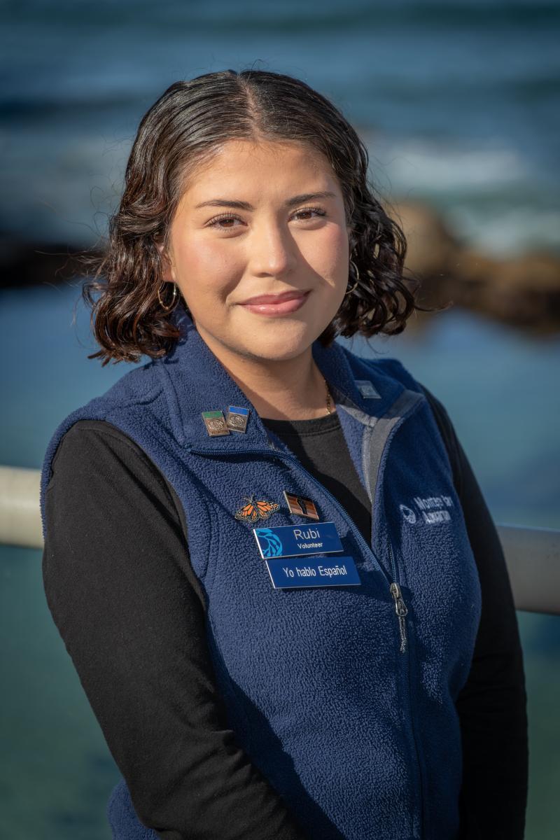 A teen volunteer wearing a blue vest smiles against an ocean background