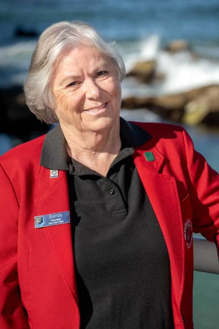 A woman with white hair wearing a red jacket smiles in front of an ocean background