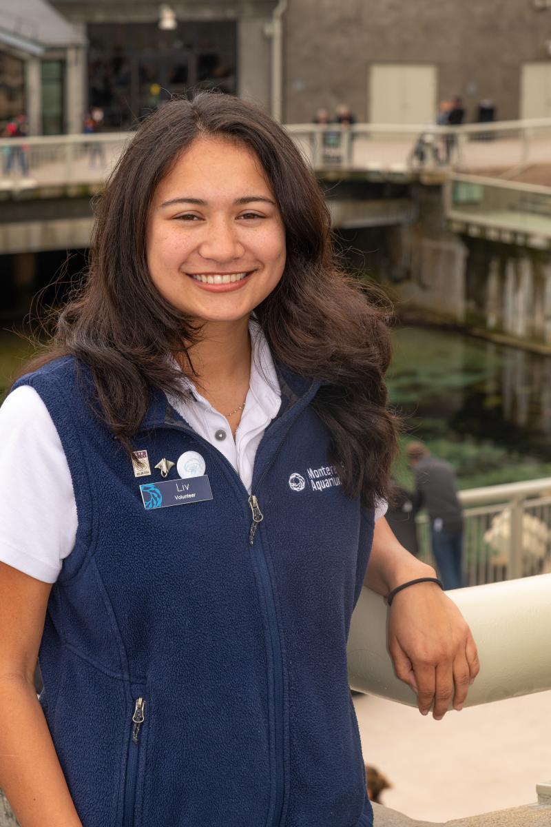 A teen volunteer in a blue vest smiles while standing on the back deck of the Aquarium