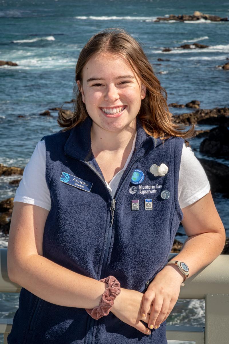 A teen volunteer in a blue vest smiles while standing in front of an ocean background