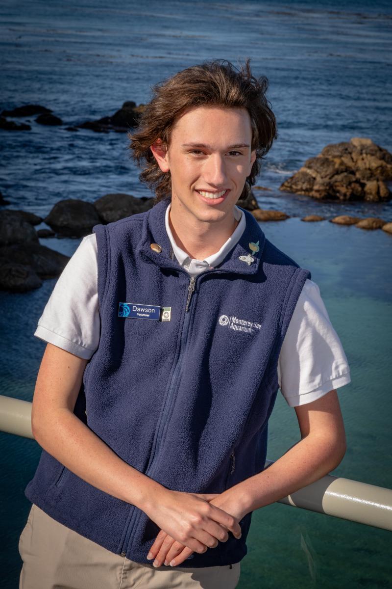 A teen volunteer in a blue vest smiles while standing in front of an ocean background