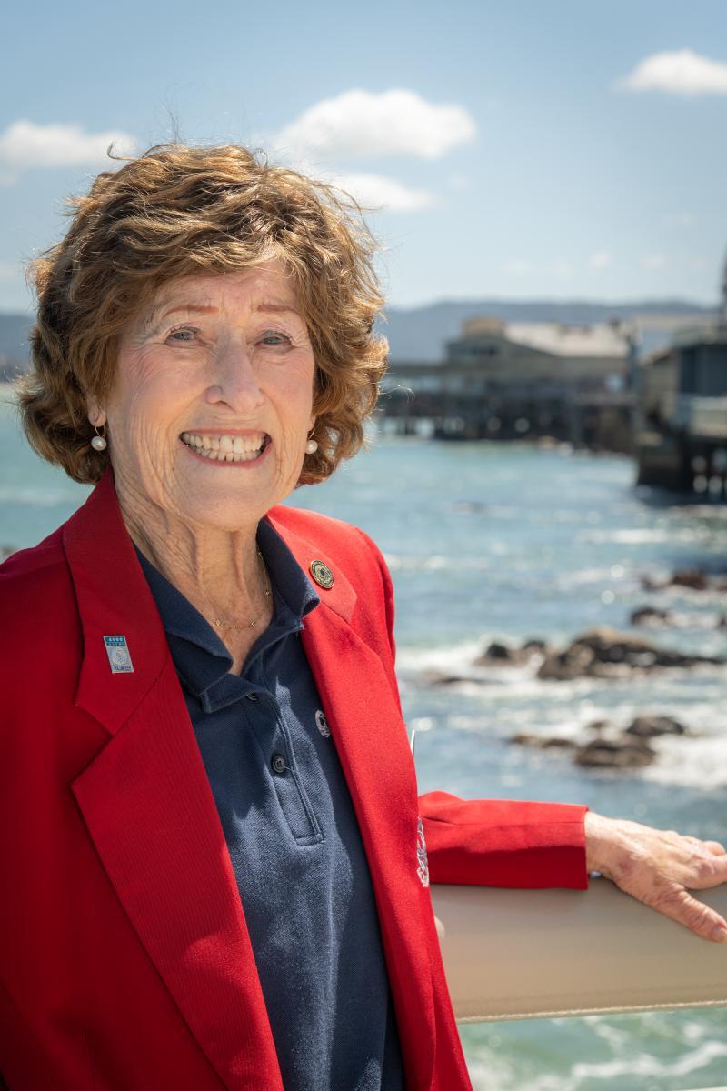 A volunteer in a red sports jacket smiles while standing on the back deck of the Aquarium