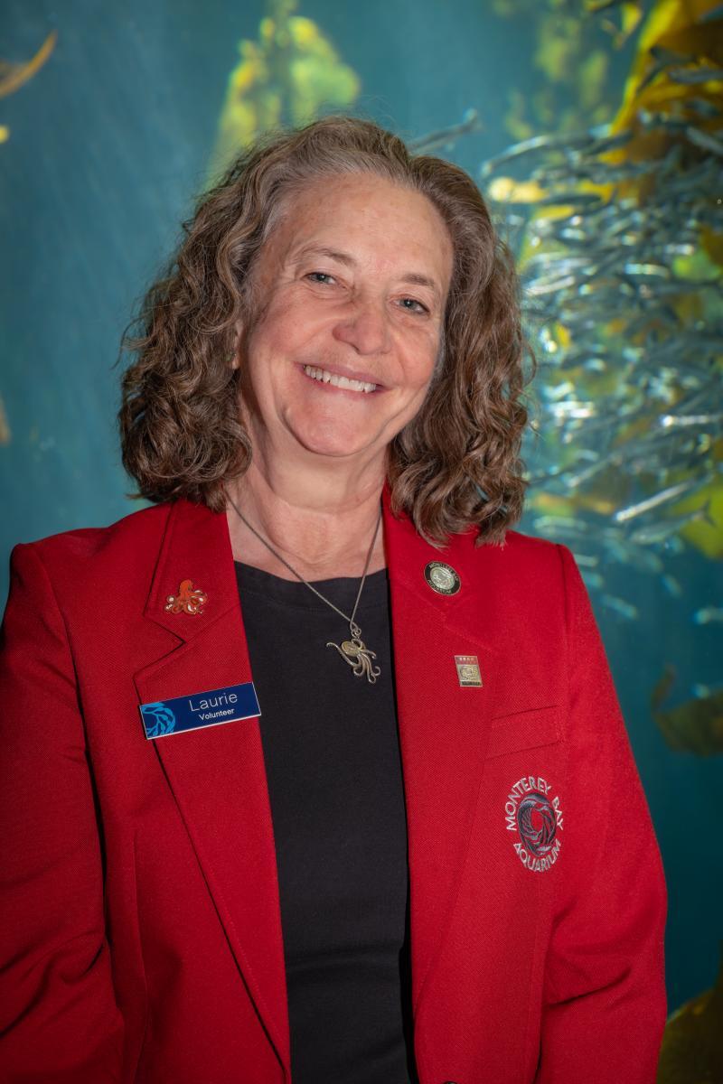 A volunteer in a red sports jacket smiles while standing in front of the Kelp Forest exhibit at the Aquarium