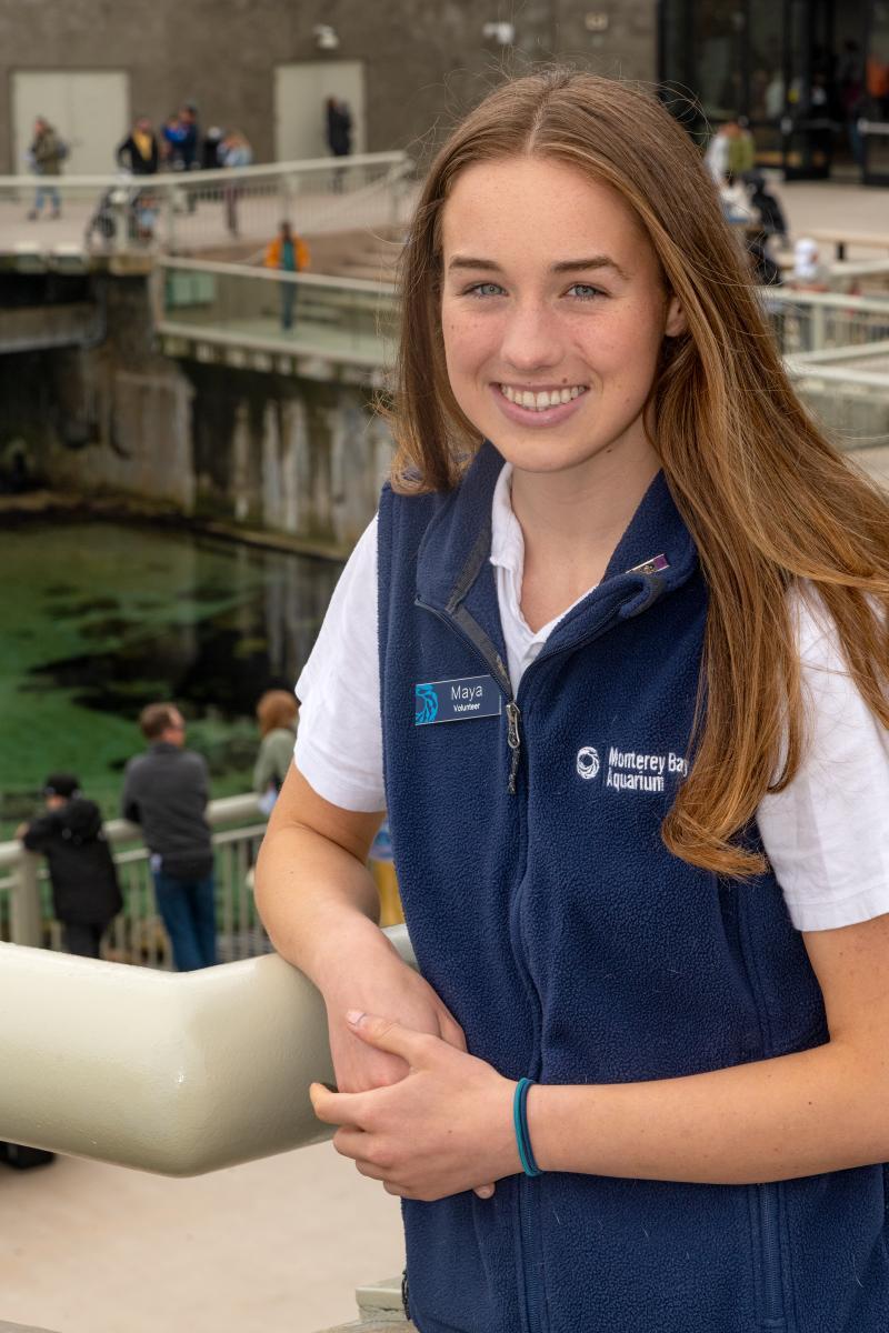 A teen volunteer in a blue vest smiles while standing on the back deck of the Aquarium