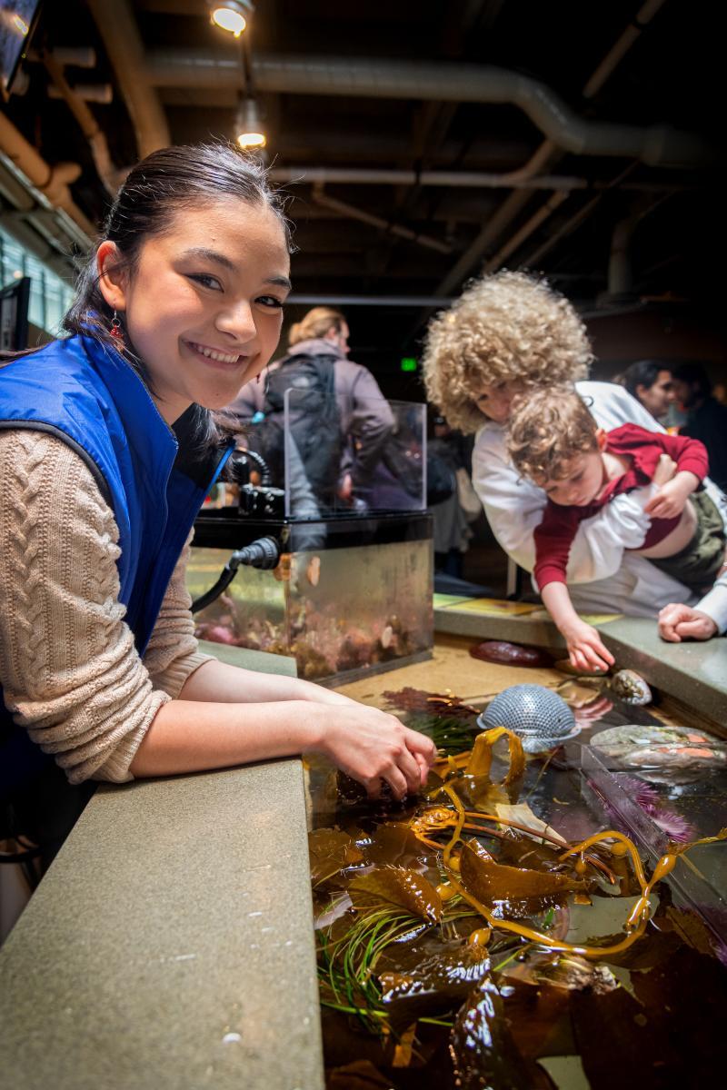 A teen volunteer in a blue vest smiles while leaning over the touch pools at the Aquarium