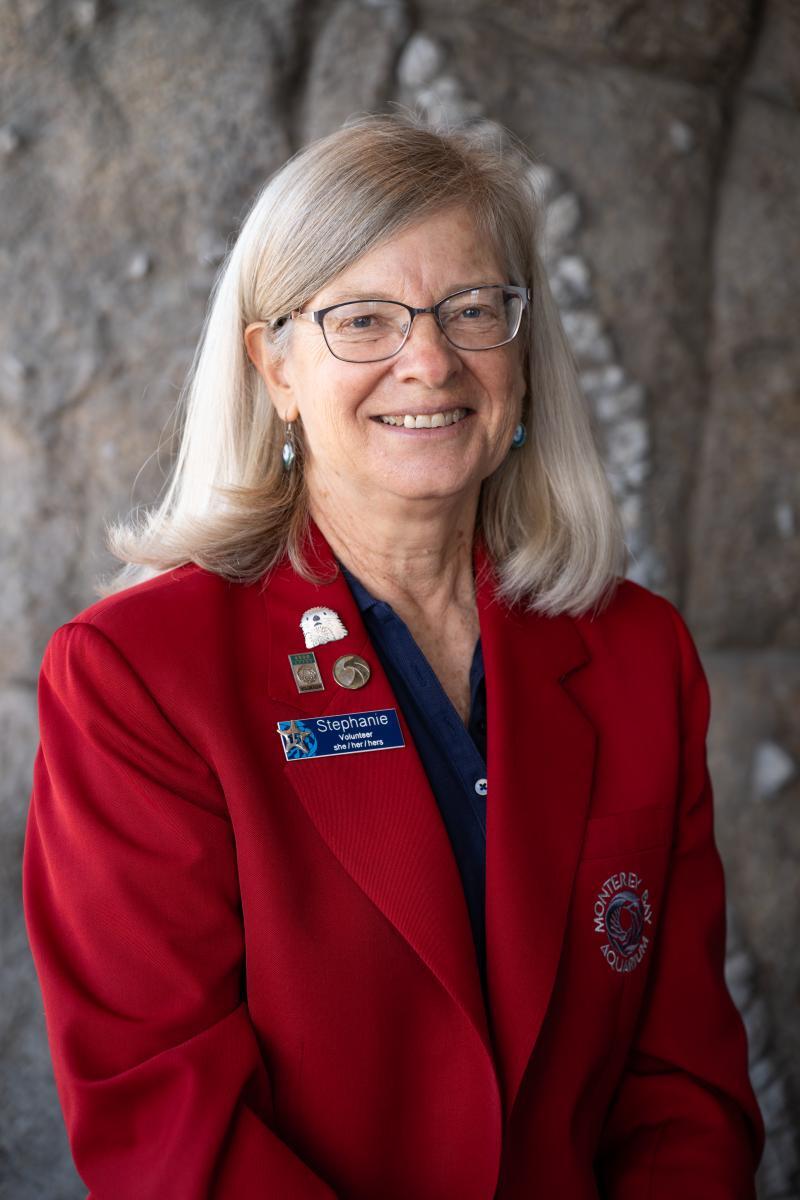 A volunteer in a red sports jacket smiles while standing in front of a sculptured wall at the Aquarium