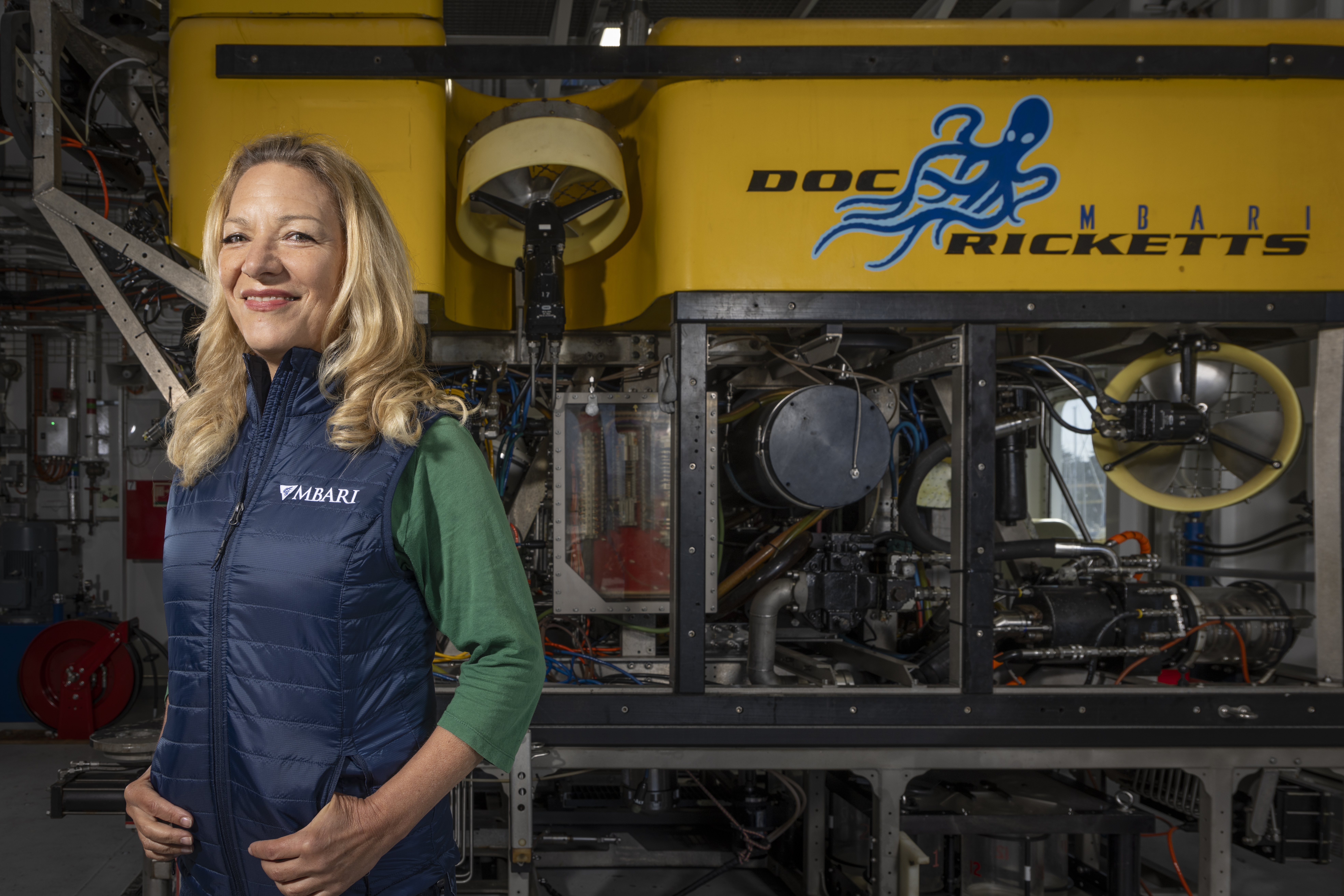 A woman with blonde hair stands smiling next to a large yellow robotic submersible.