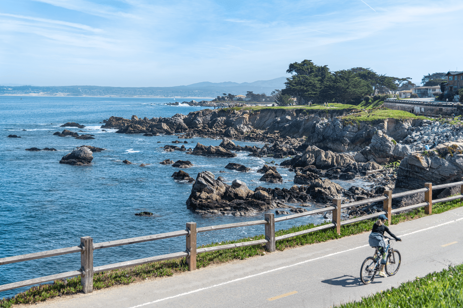 A cyclist rides a coastal path beside a wooden rail fence overlooking rocky shores and blue ocean