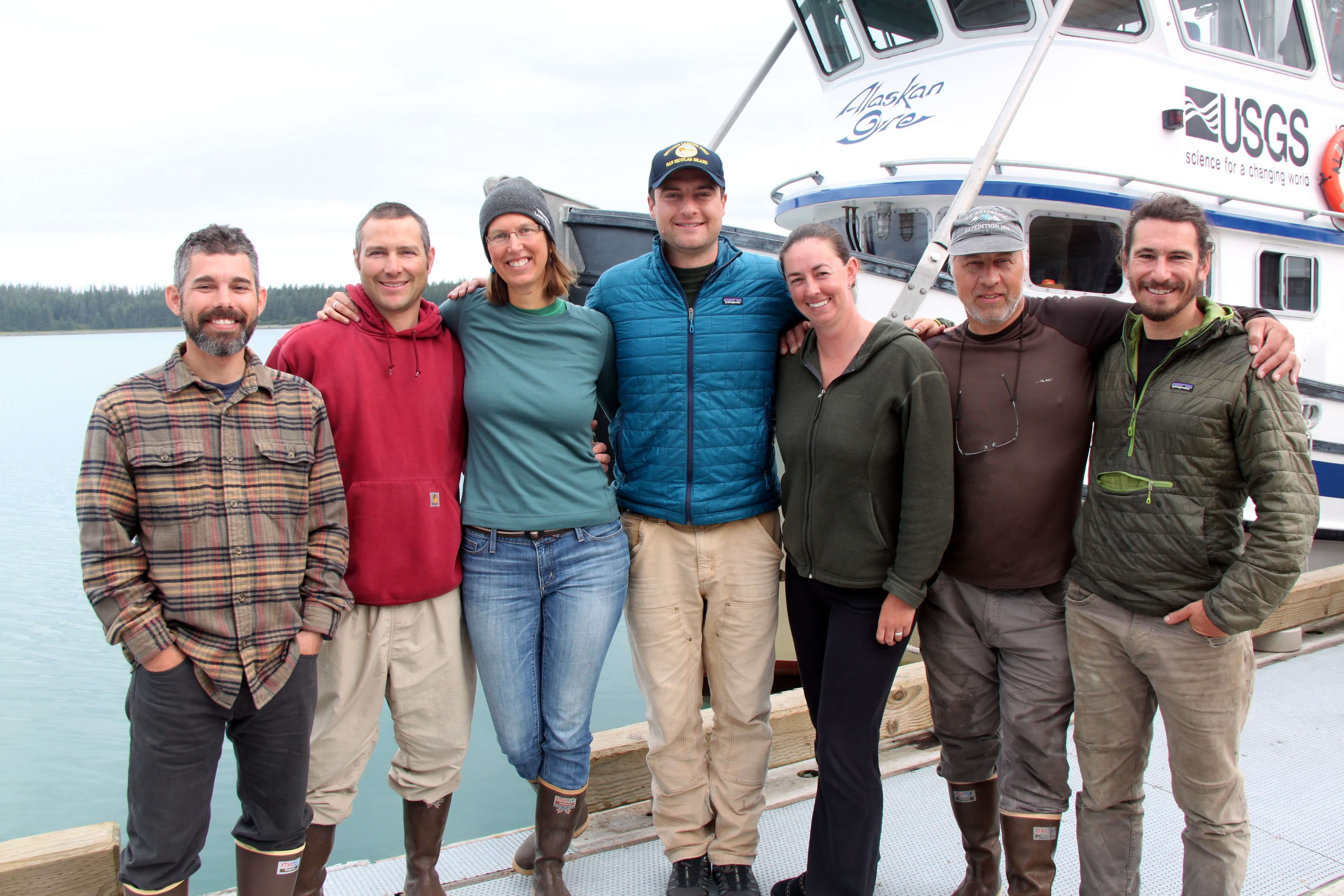 Jess posing shoulder to shoulder with other researchers in front of the glassy calm waters of Glacier Bay and the USGS research vessel the Alaskan Gyre