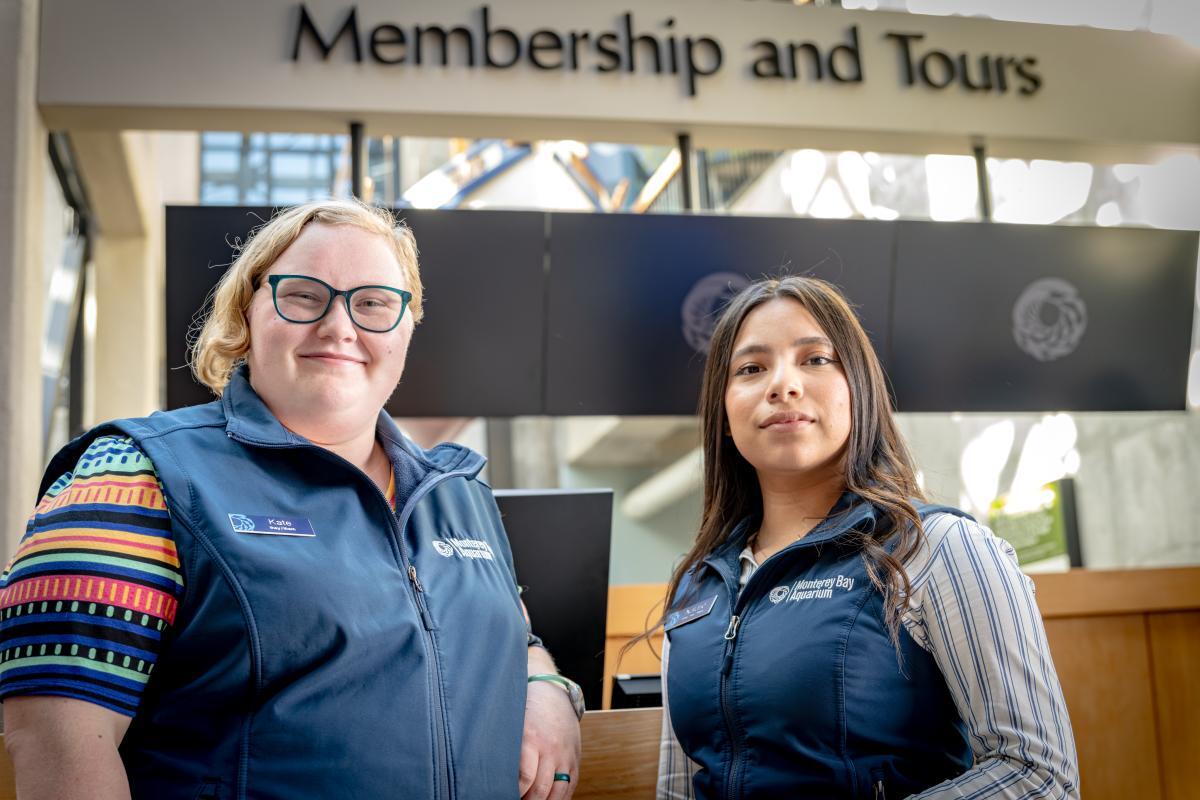 Two people wearing blue Aquarium vests stand in front of a sign that reads Membership and Tours