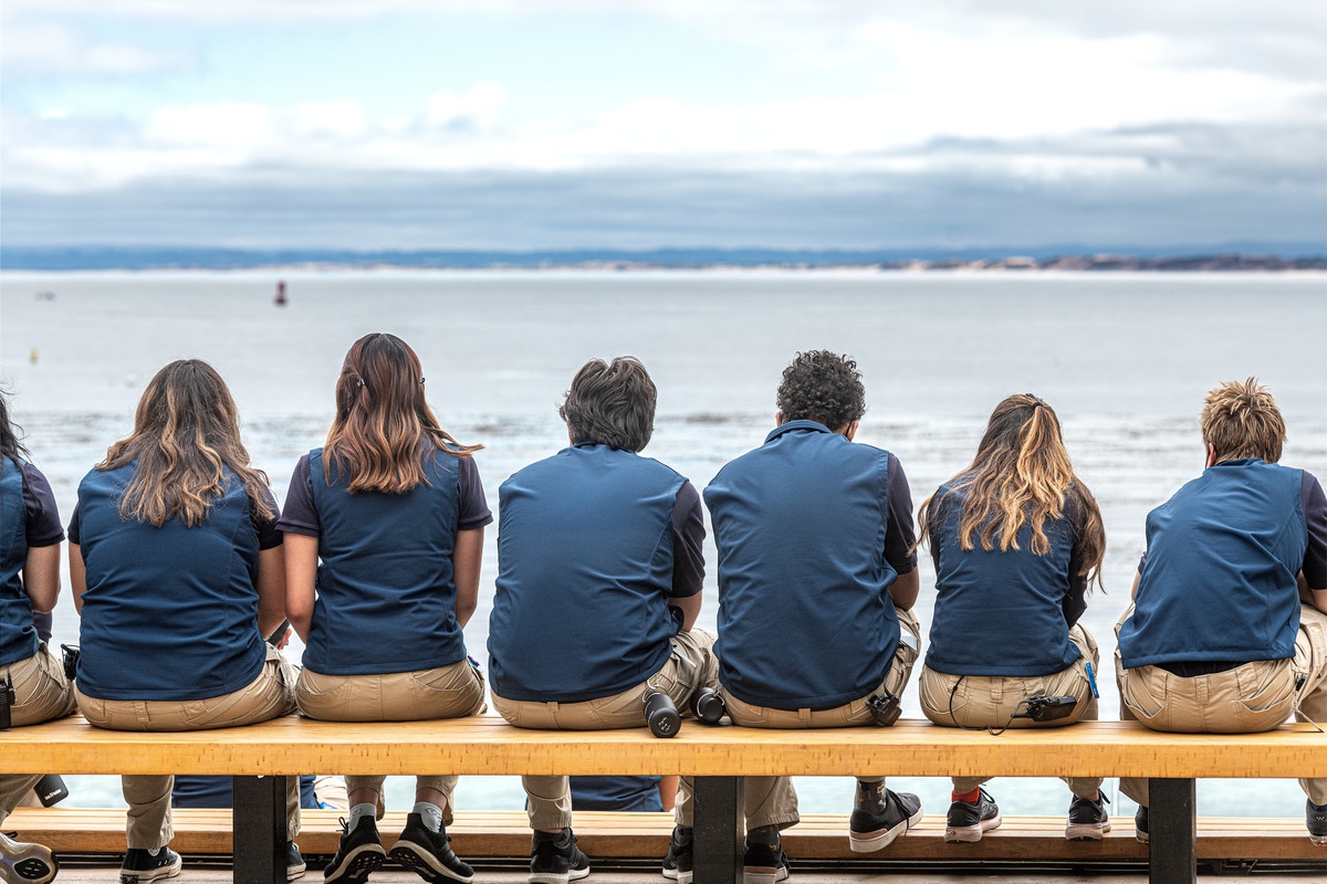 Seven uniformed Aquarium staff members sitting on a bench on the back deck with their backs towards the ocean