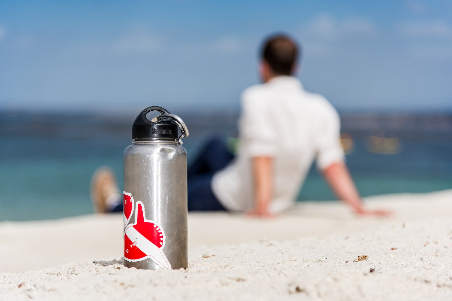 Reusable metal water bottle with a sticker on a sandy beach, person sitting by the ocean in the background