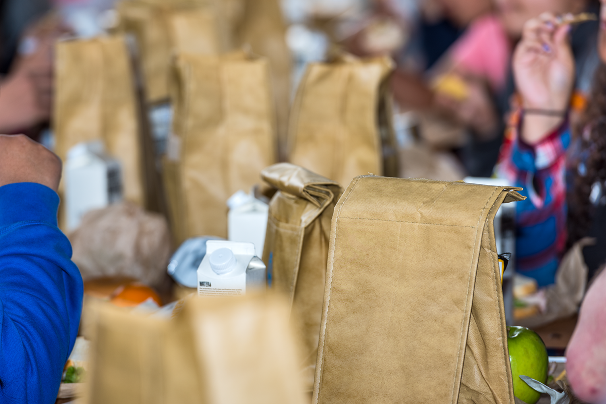 Students at lunch tables with brown paper bag meals and milk cartons