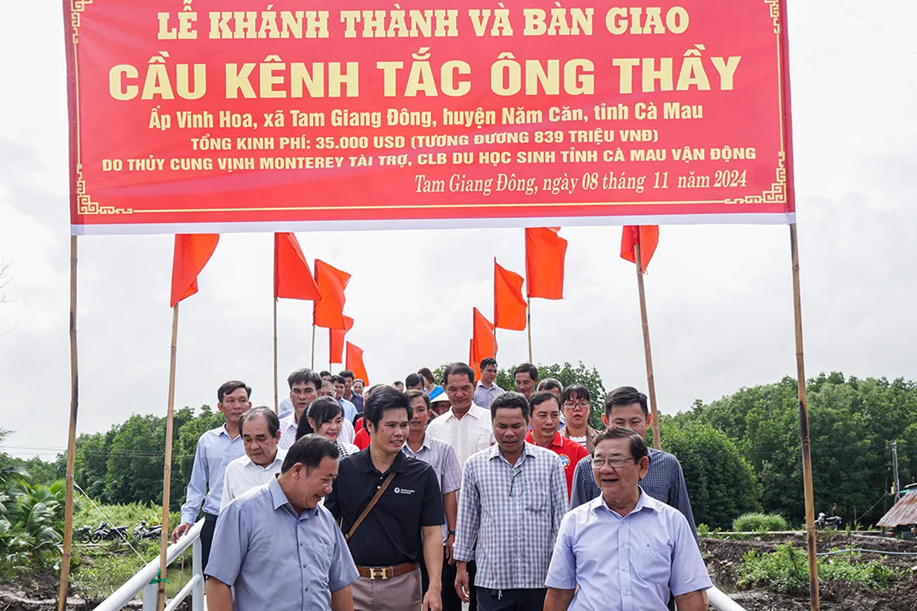 Officials and community members walking across the newly inaugurated Kenh Tac Ong Thay bridge in Ca Mau, Vietnam, with a ceremonial red banner overhead marking the dedication event