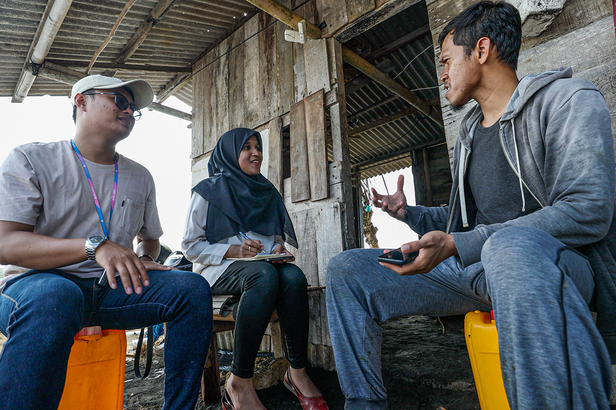 Field researchers interviewing a community member outside a weathered coastal structure, with one taking notes and another listening attentively