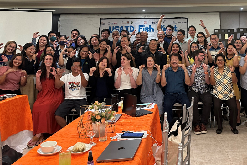 Large group of USAID Fish Right program participants posing and smiling together at a workshop, making fish-shaped hand gestures in front of a project banner