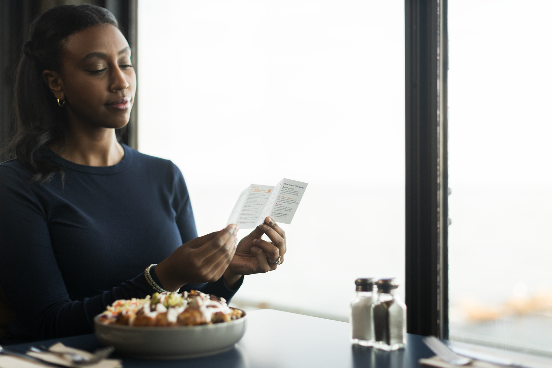 Person reads a Seafood Watch pocket guide at a restaurant table with a food bowl in front of her