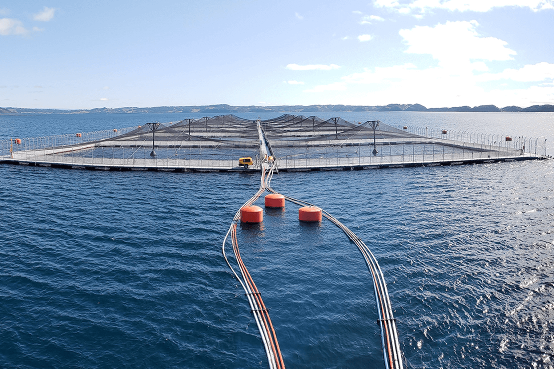 An aerial view of a large open-ocean salmon farm with net pen structures and orange buoys in blue water
