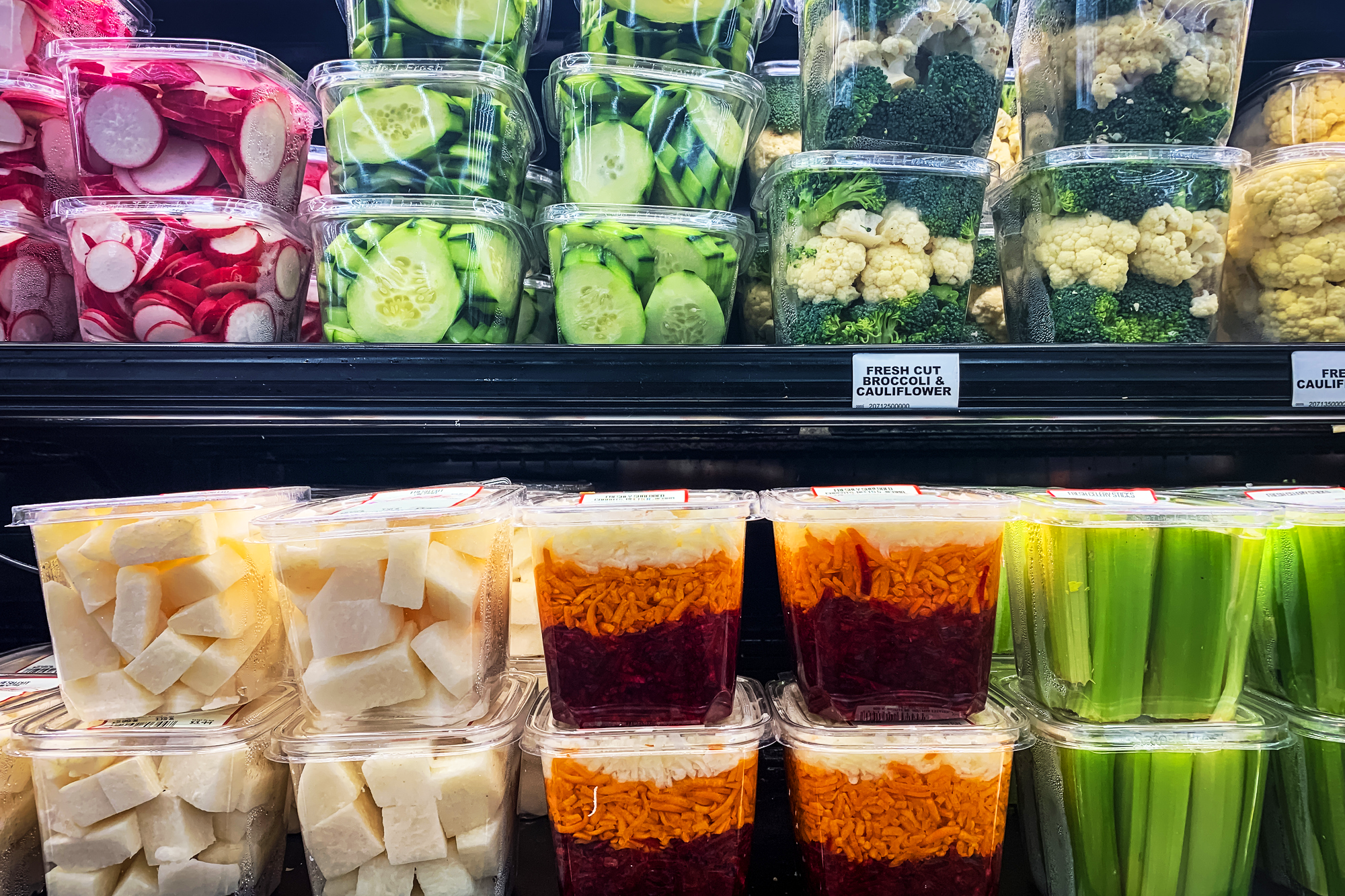 Close-up of a refrigerated grocery shelf stocked with plastic containers of vegetables, including sliced radish, cucumber, broccoli, cauliflower, celery, and shredded carrot