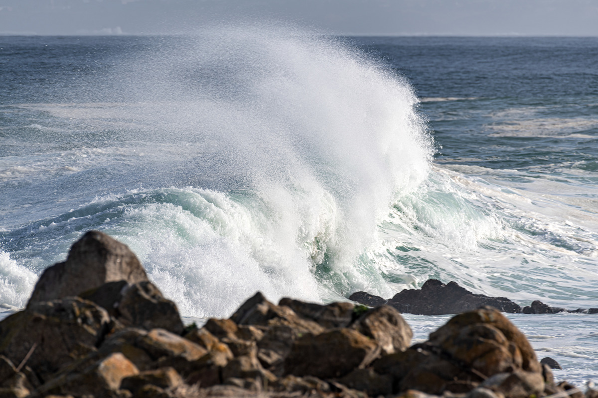 A large ocean wave breaks onto a small island of rocks, tossing a spray of water behind as it crashes