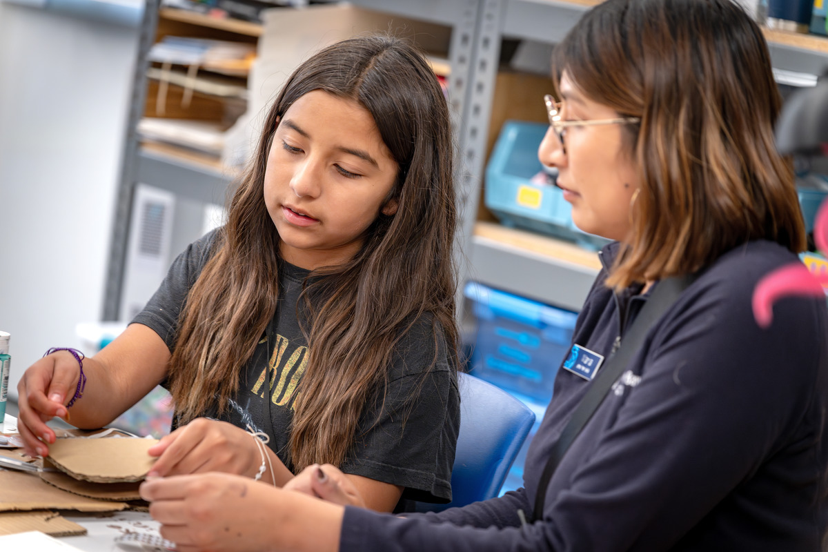 A middle school student with long brown hair looks down at her cardboard project while an Aquarium staff member with shorter brown hair sits nearby looking on