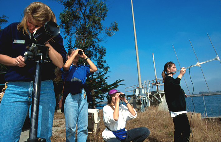 Four women spotting sea otters while holding various pieces of equipment.